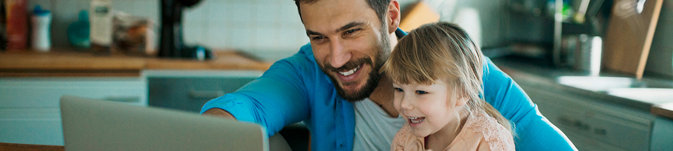 Father sitting at kitchen table with young daughter on his lap pointing at his laptop and smiling. 